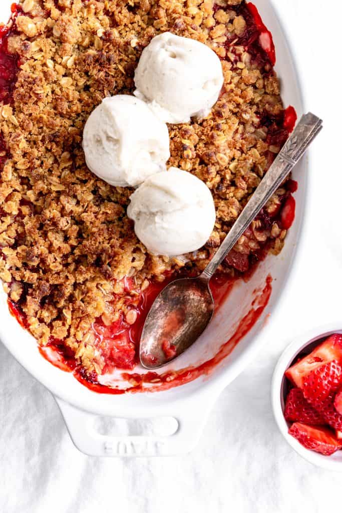 Spoon digging into a strawberry crisp topped with scoops of vanilla ice cream.