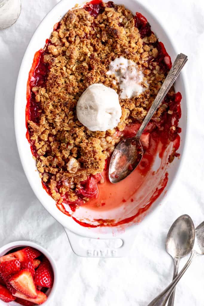 Spoons digging into a dish of vegan strawberry crisp and a cup of strawberries.