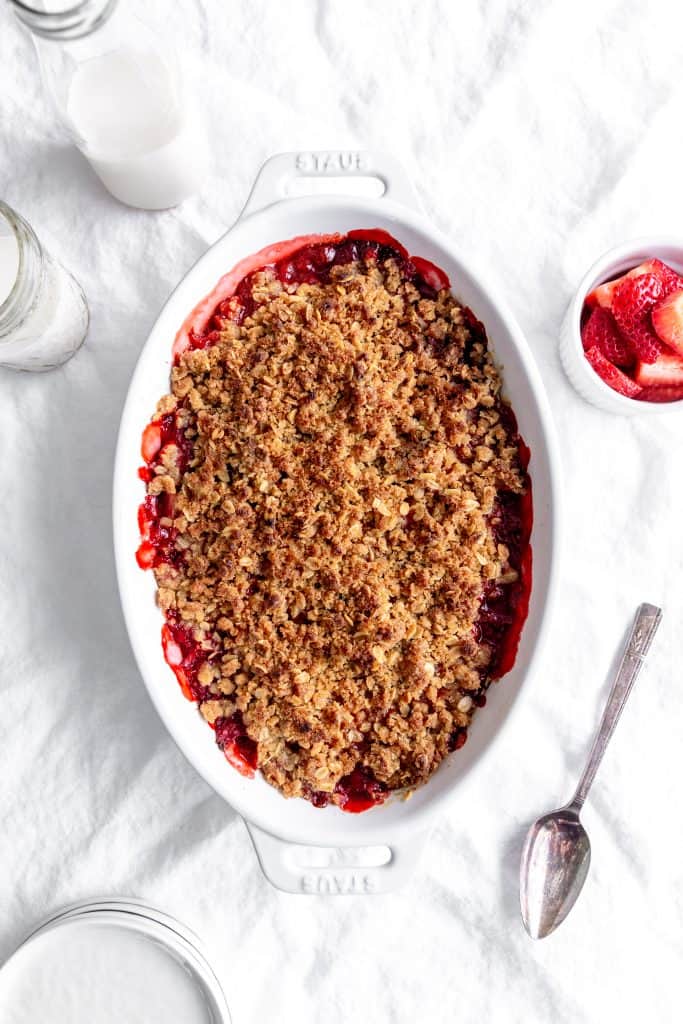 Strawberry crisp in a white baking dish, a cup of strawberries and a jug of milk.