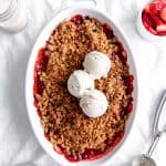 Baking dish of strawberry crisp topped with scoops of vanilla ice cream and a jug of milk.