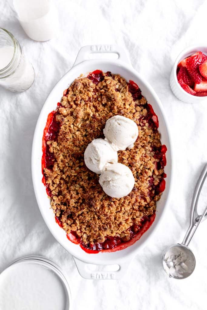 Baking dish of strawberry crisp topped with scoops of vanilla ice cream and a jug of milk.