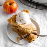 Fork cutting into slice of apple pie topped with vanilla ice cream.