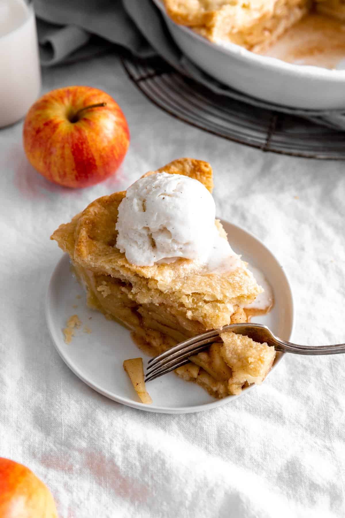 Fork cutting into slice of apple pie topped with vanilla ice cream.