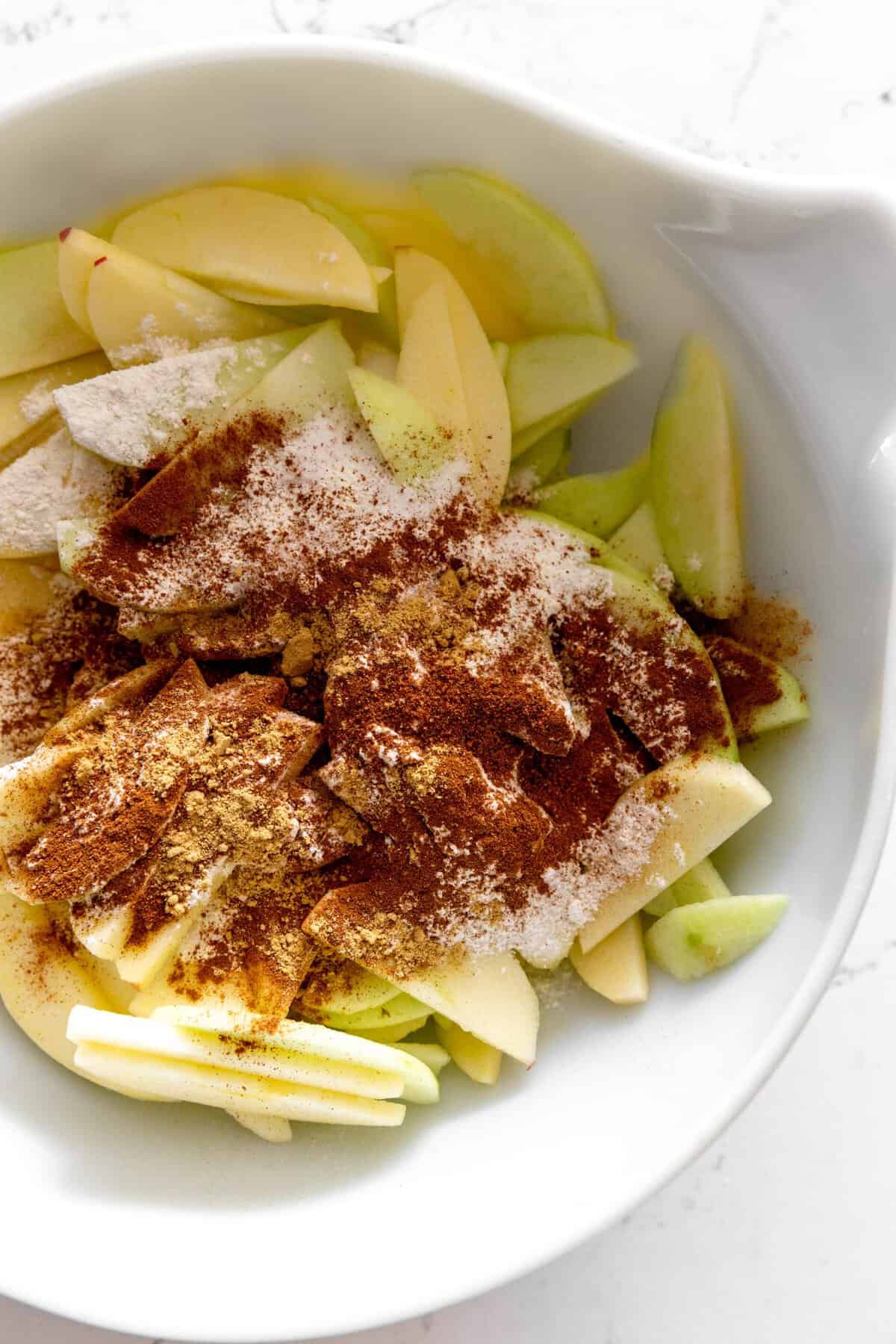 Sliced apples covered in sugar in spices in a white bowl.