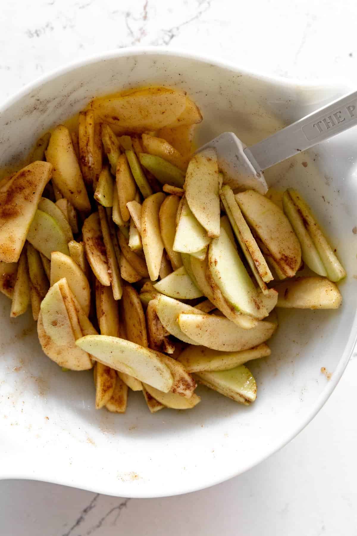 Sliced apples tossed with sugar and spices in a white bowl with a rubber spatula.