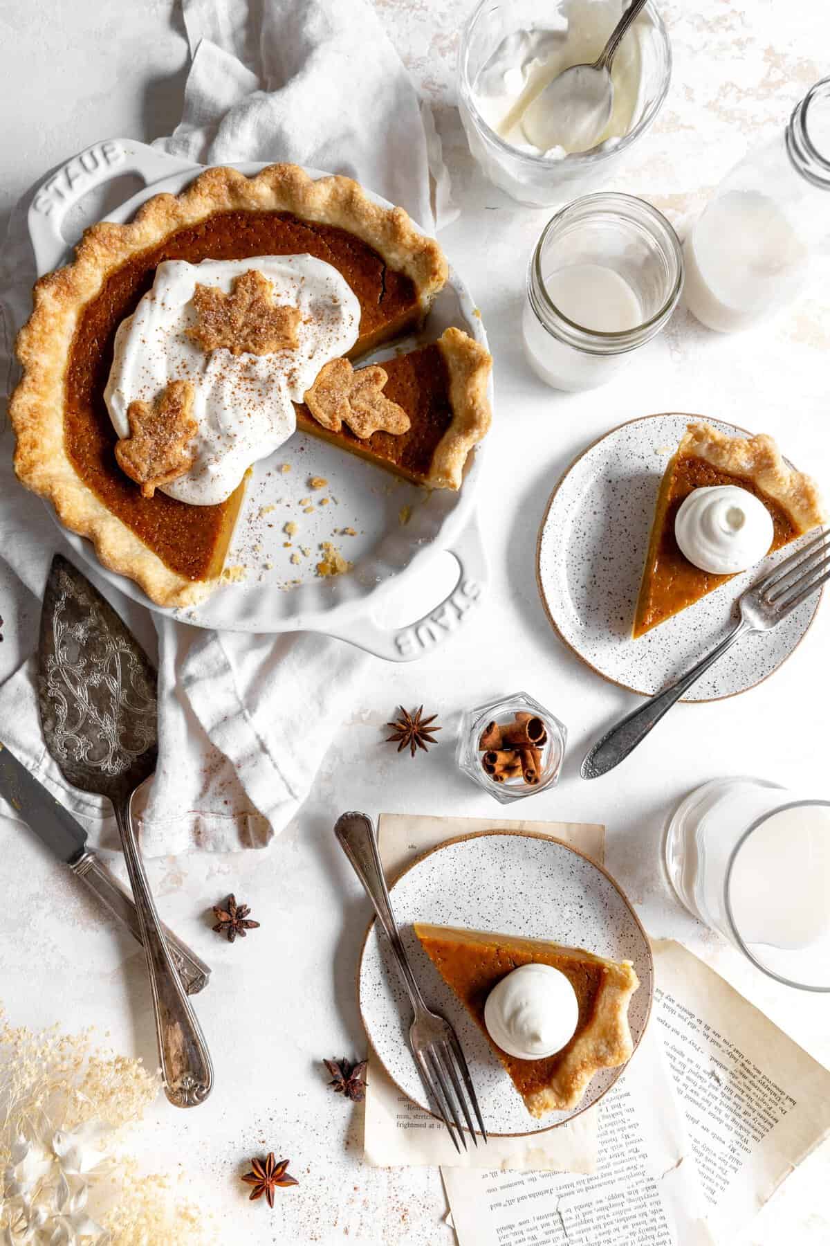 Pumpkin pie, slices of pumpkin pie on plates, a jug of milk, cutlery and cinnamon sticks.