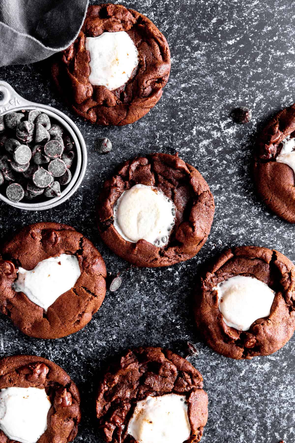 Hot chocolate cookies on a black surface with a dark linen napkin and a cup of chocolate chips.