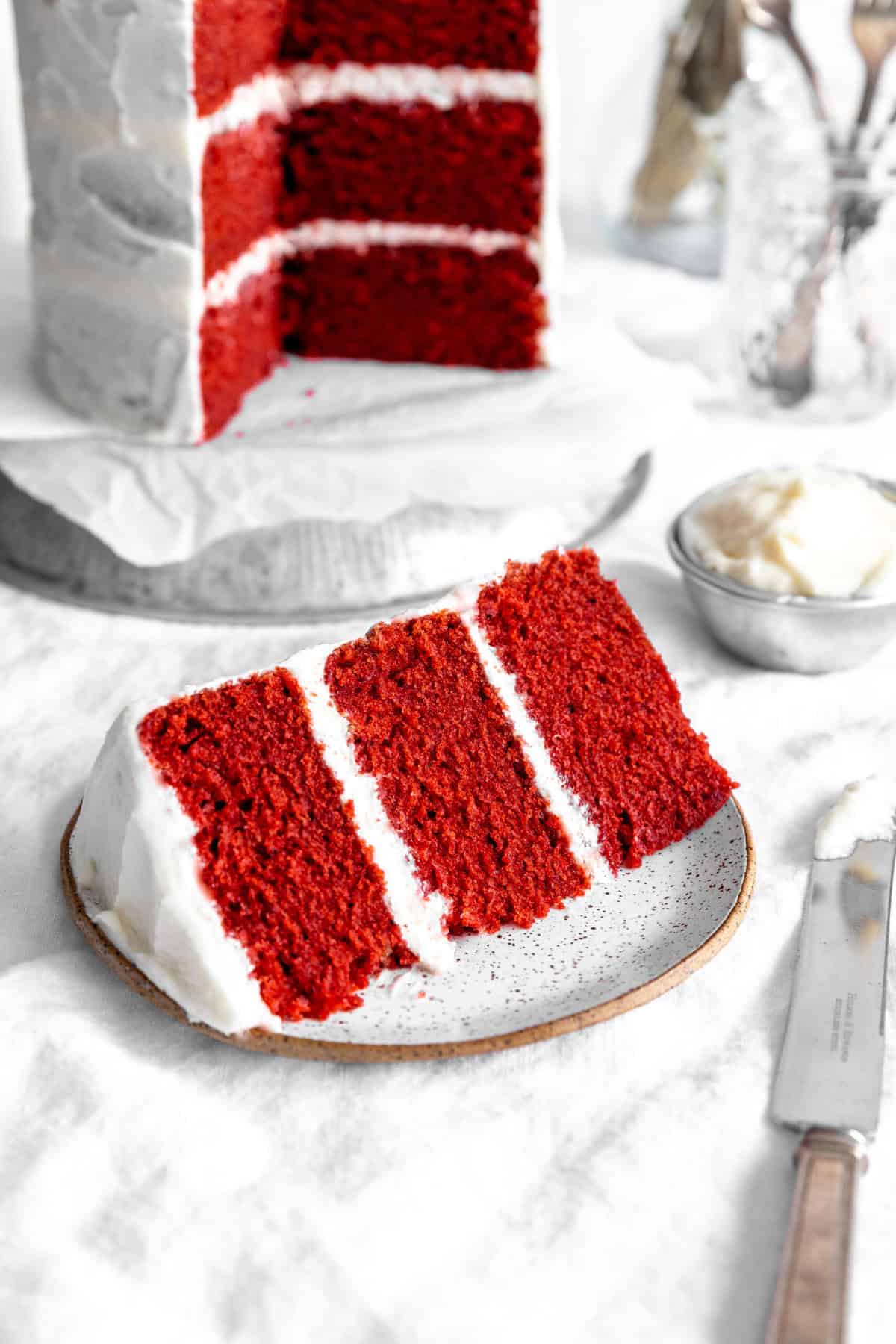 Slice of red velvet cake on a white plate in front of the whole layer cake.