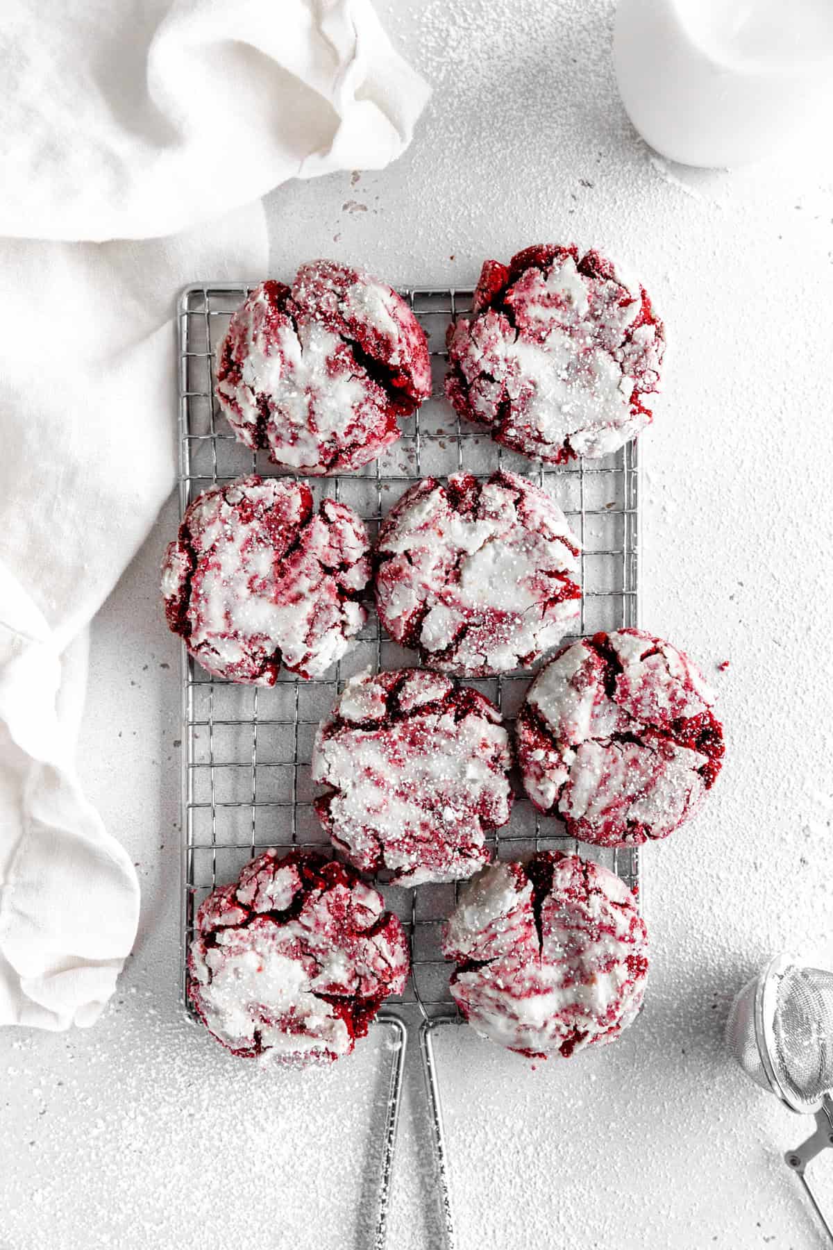 Red velvet crinkle cookies on a wire cooling rack, a linen napkin and a jug of milk.