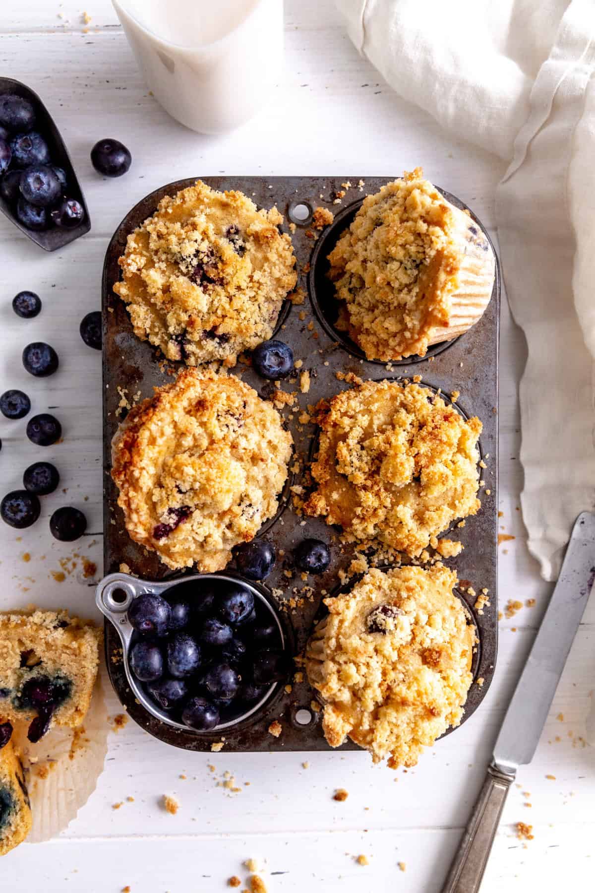 Pan of blueberry muffins, a cup of fresh blueberries, a knife and a cup of milk.