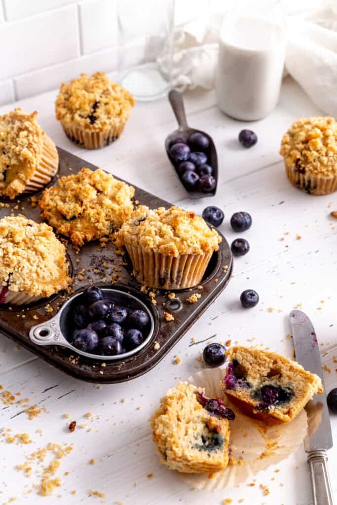 Blueberry muffins in a baking pan, a knife, fresh blueberries and a jug of milk.