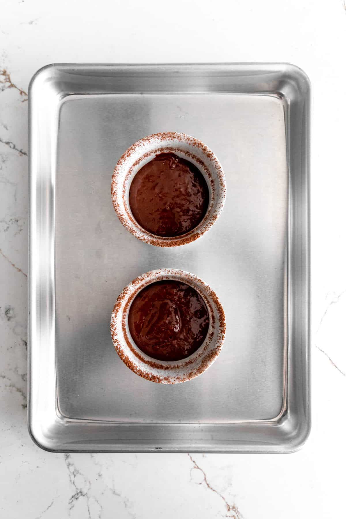 Chocolate cake batter in two white ramekins on a baking sheet.