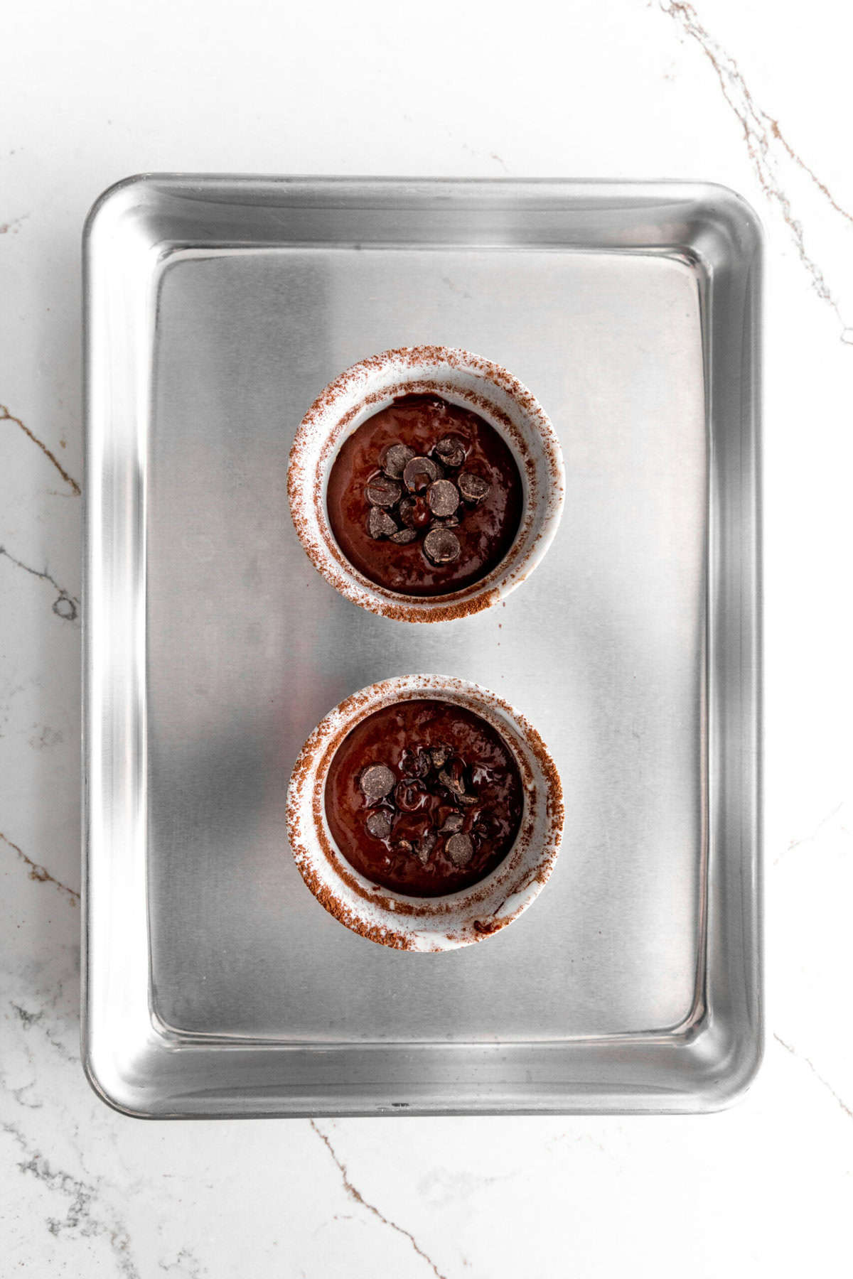 Chocolate cake batter topped with chocolate chips in two white ramekins on a baking sheet.