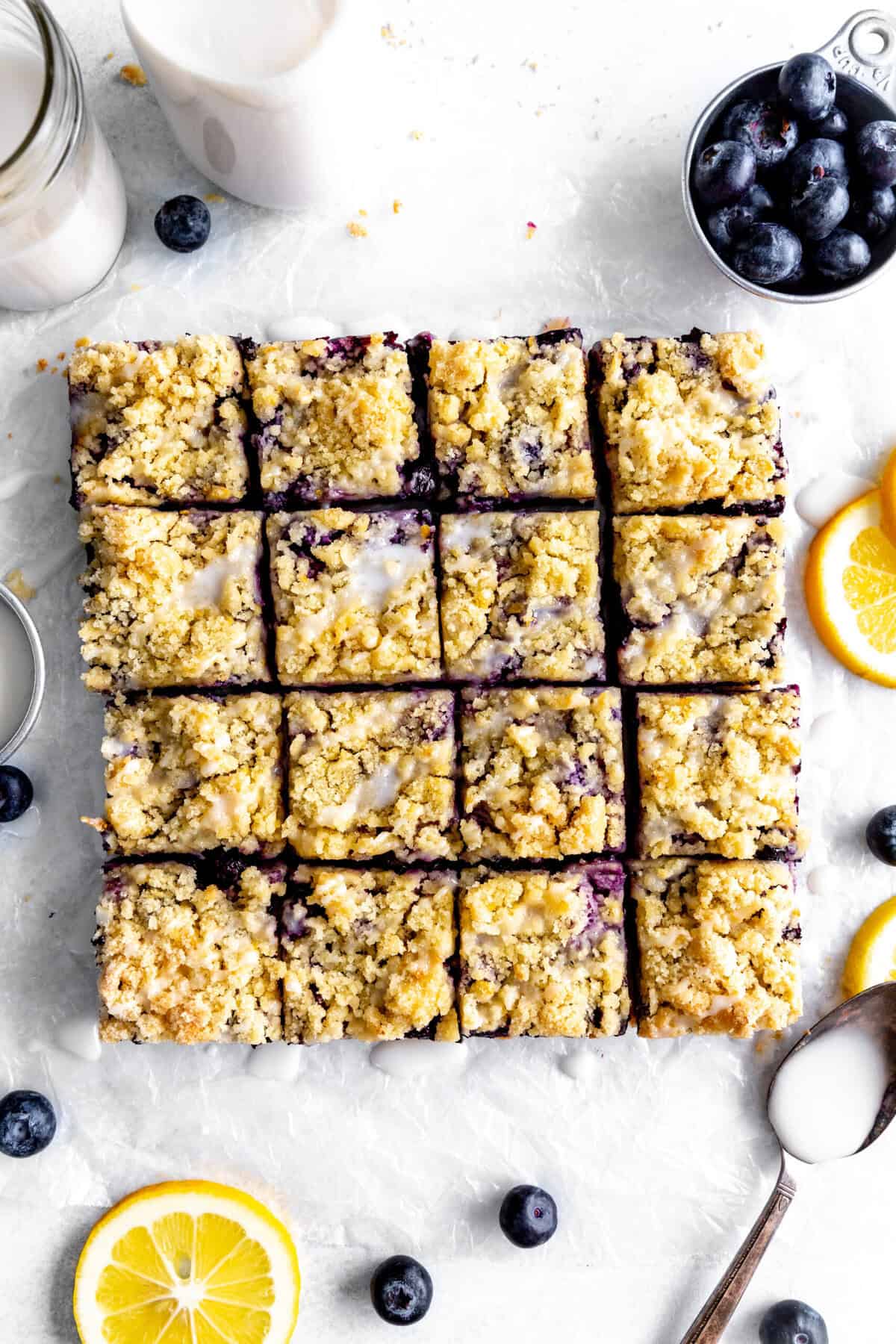 Lemon blueberry bars arranged in a square, lemon slices, a cup of blueberries and a cup of milk.