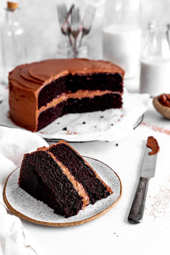 Slice of chocolate cake on a plate in front of the rest of the cake and a jug of milk.