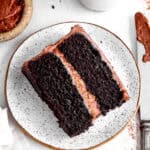 Slice of chocolate cake on a white plate, a jug of milk and a knife covered in frosting.