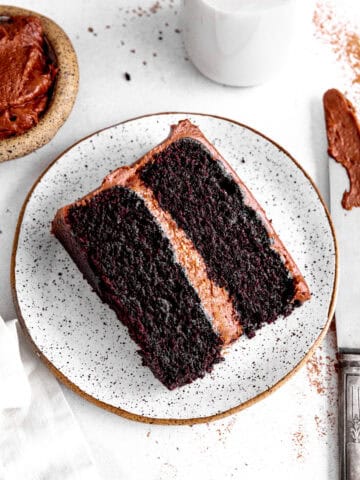 Slice of chocolate cake on a white plate, a jug of milk and a knife covered in frosting.