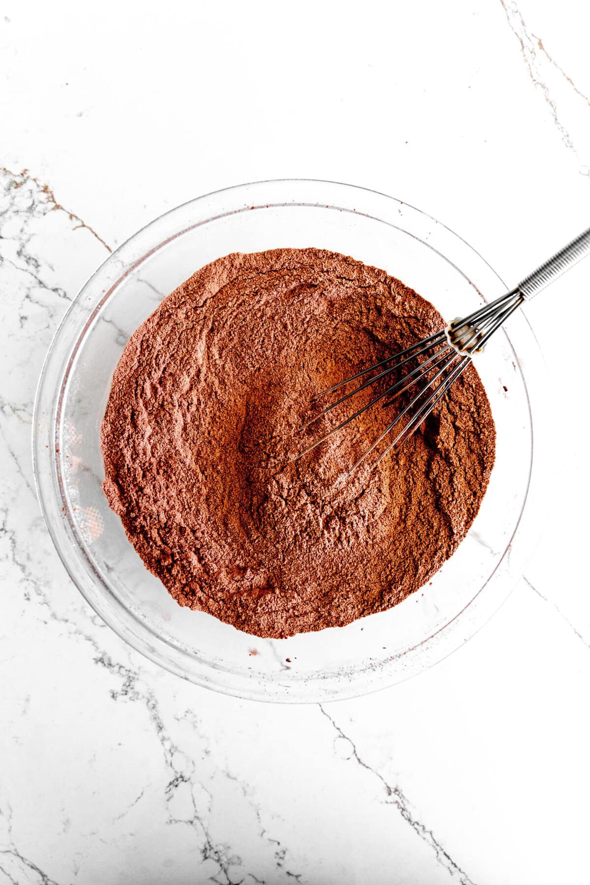 Glass bowl filled with dry chocolate cake ingredients and a wire whisk.