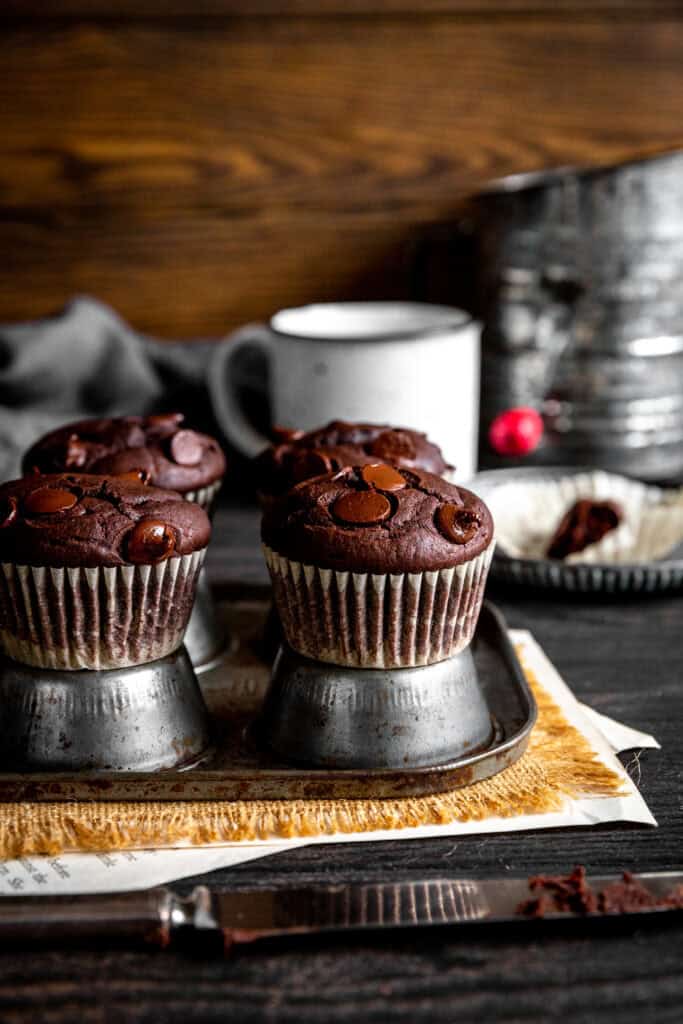 Chocolate muffins on an upside down muffin pan, a sugar sifter and a white mug.