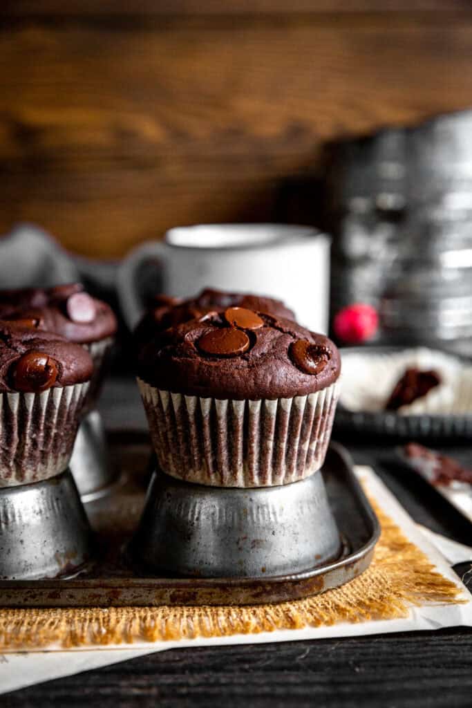 Chocolate muffins on an upside down muffin pan, a sugar sifter and a white mug.