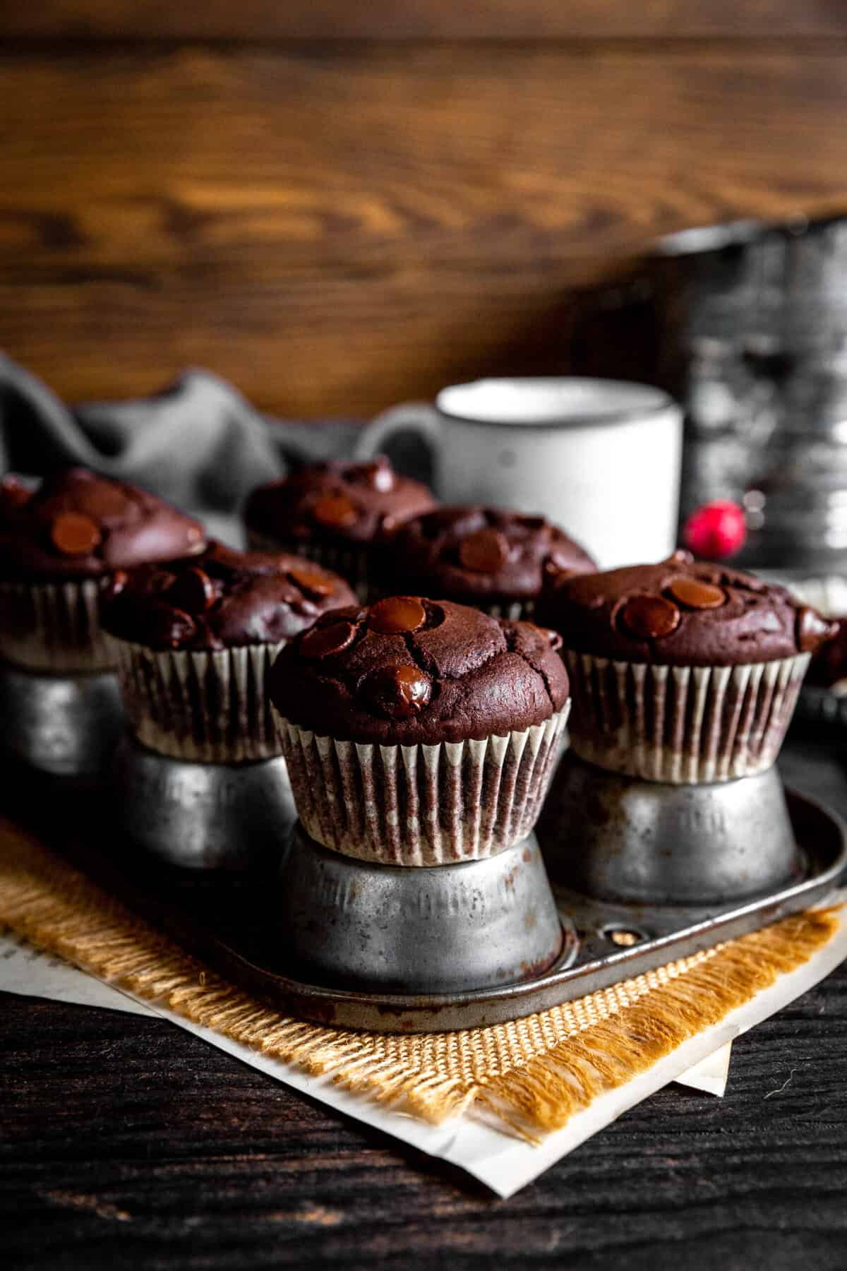 Chocolate muffins on an upside down muffin pan, a sugar sifter and a white mug.