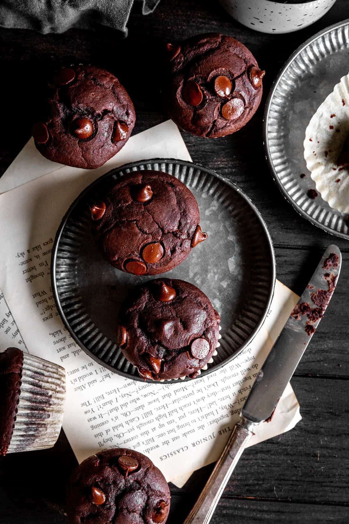 Chocolate muffins on metal plates, a knife and a mug all on a dark wood surface.