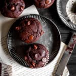 Chocolate muffins on metal plates, a knife and a mug all on a dark wood surface.