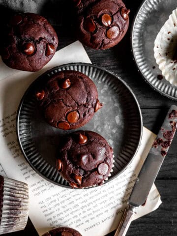 Chocolate muffins on metal plates, a knife and a mug all on a dark wood surface.