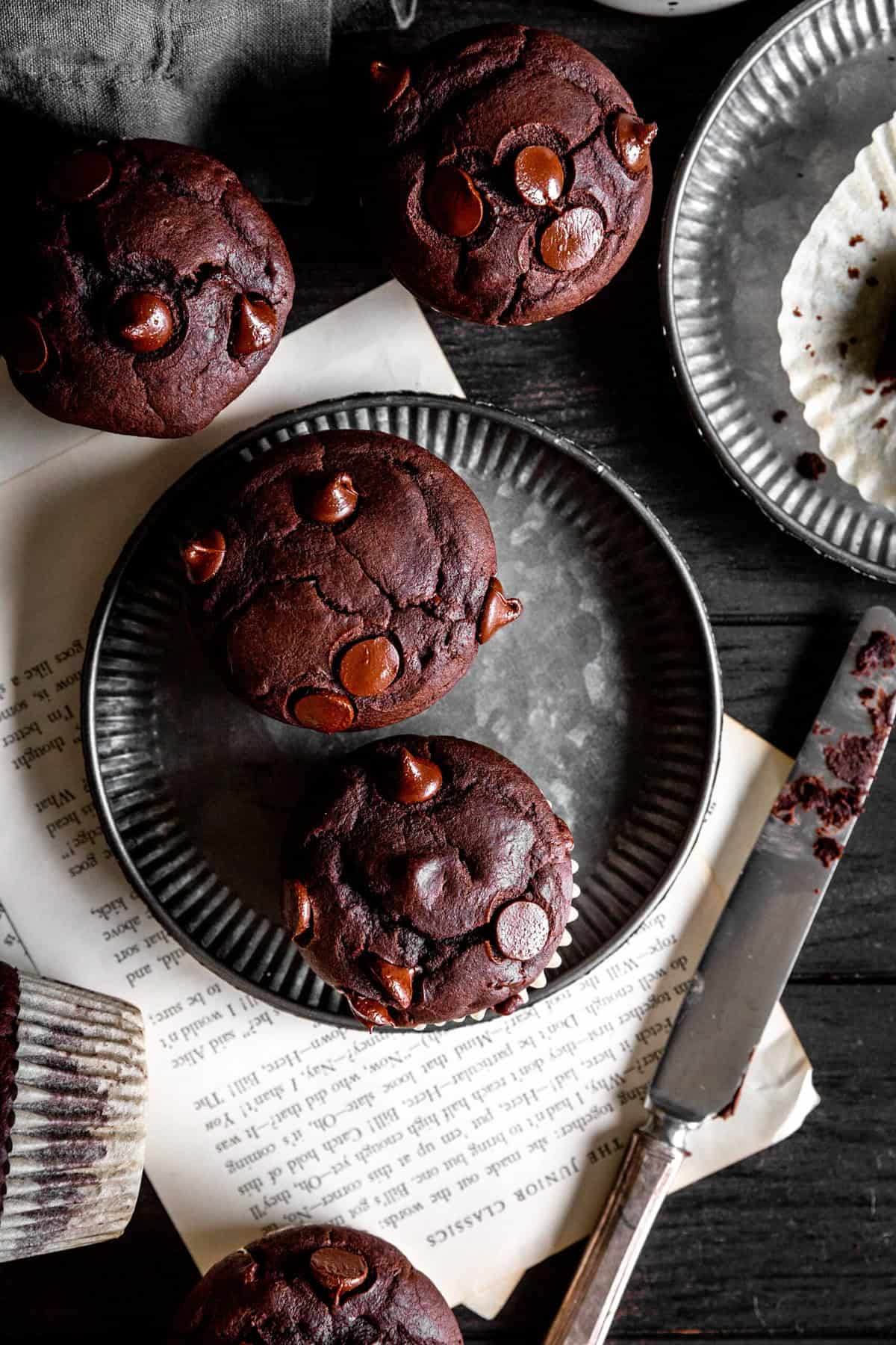 Chocolate muffins on metal plates, a knife and a mug all on a dark wood surface.