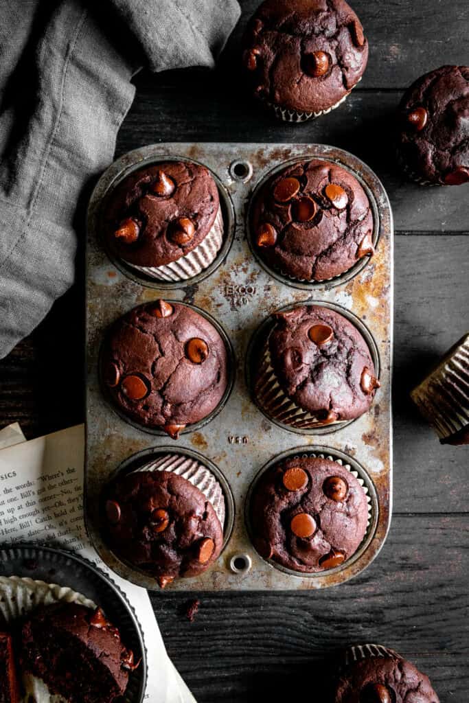 Chocolate muffins in a cupcakes pan and a gray linen napkin on a dark wood surface.