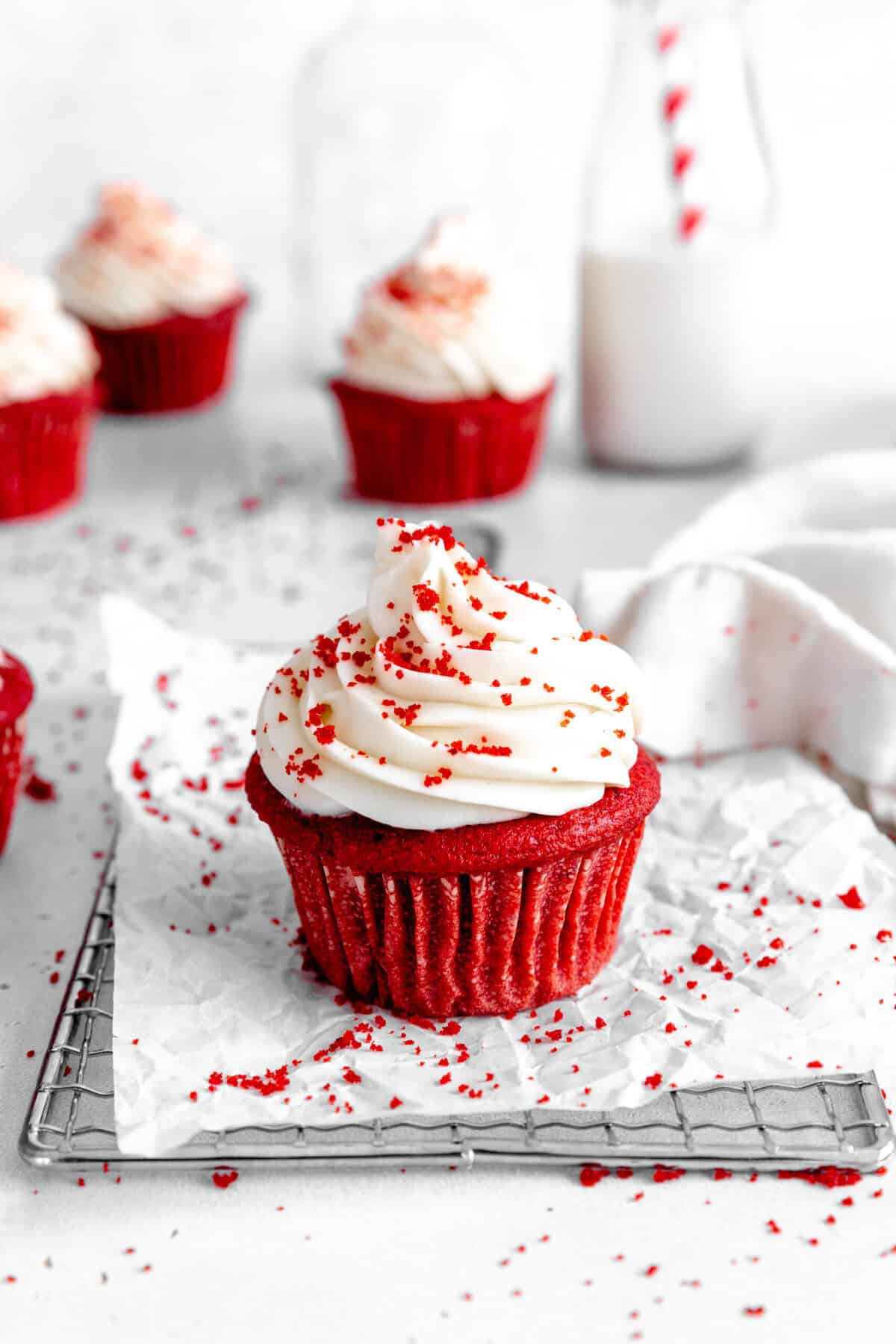 Red velvet cupcake on a wire cooling rack in front of a jug of milk and more cupcakes.