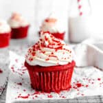 Red velvet cupcake on a wire cooling rack in front of a jug of milk and more cupcakes.