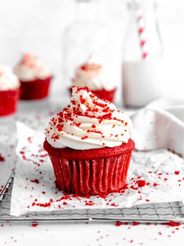 Red velvet cupcake on a wire cooling rack in front of a jug of milk and more cupcakes.