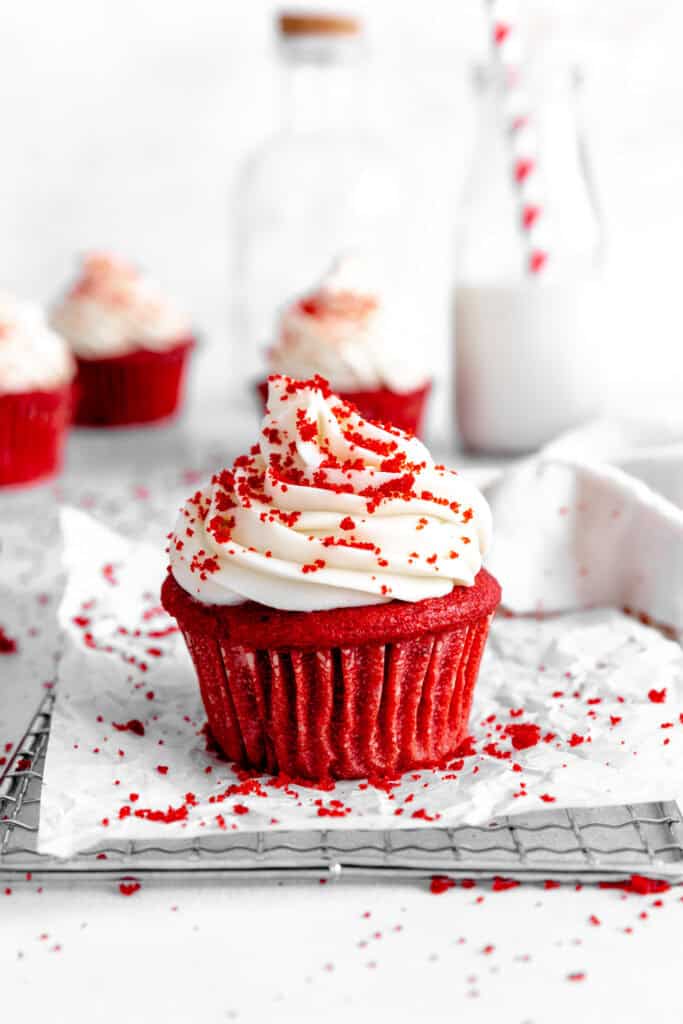 Red velvet cupcake on a wire cooling rack in front of a jug of milk and more cupcakes.