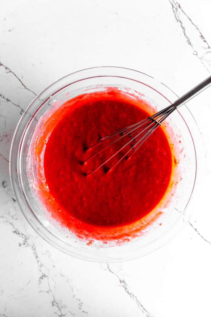 Glass bowl of red velvet cupcake batter with a wire whisk.