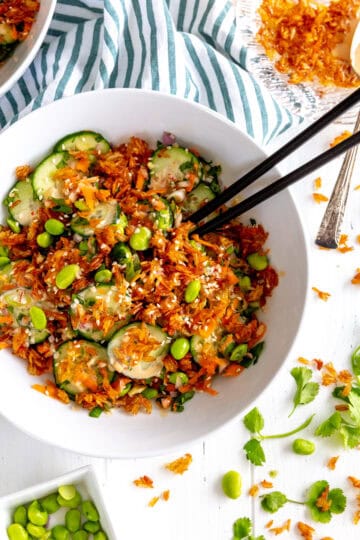 Chopsticks digging into a bowl of cucumber salad topped with crispy rice.