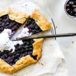 Knife cutting into a blueberry galette with a slice pulled out and a cup of milk.