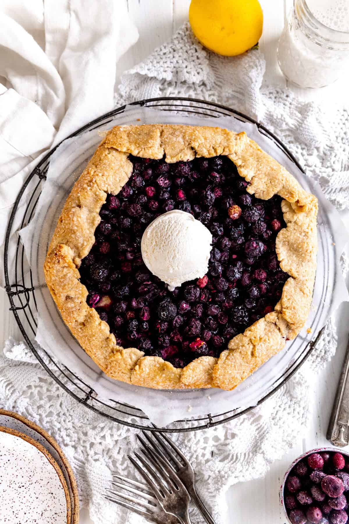 Blueberry galette on a wire rack, a cup of milk, a lemon, a stack of plates.