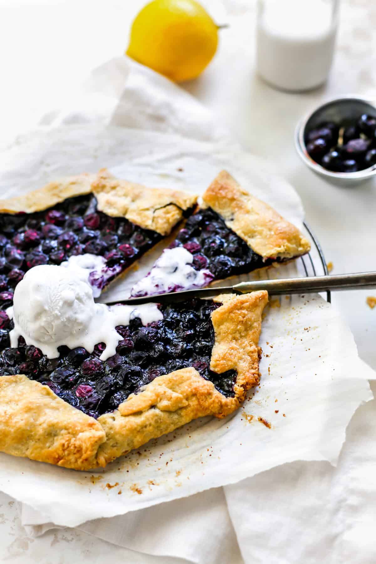 Slice being pulled out of a blueberry galette, a cup of milk, a lemon and a cup of blueberries.