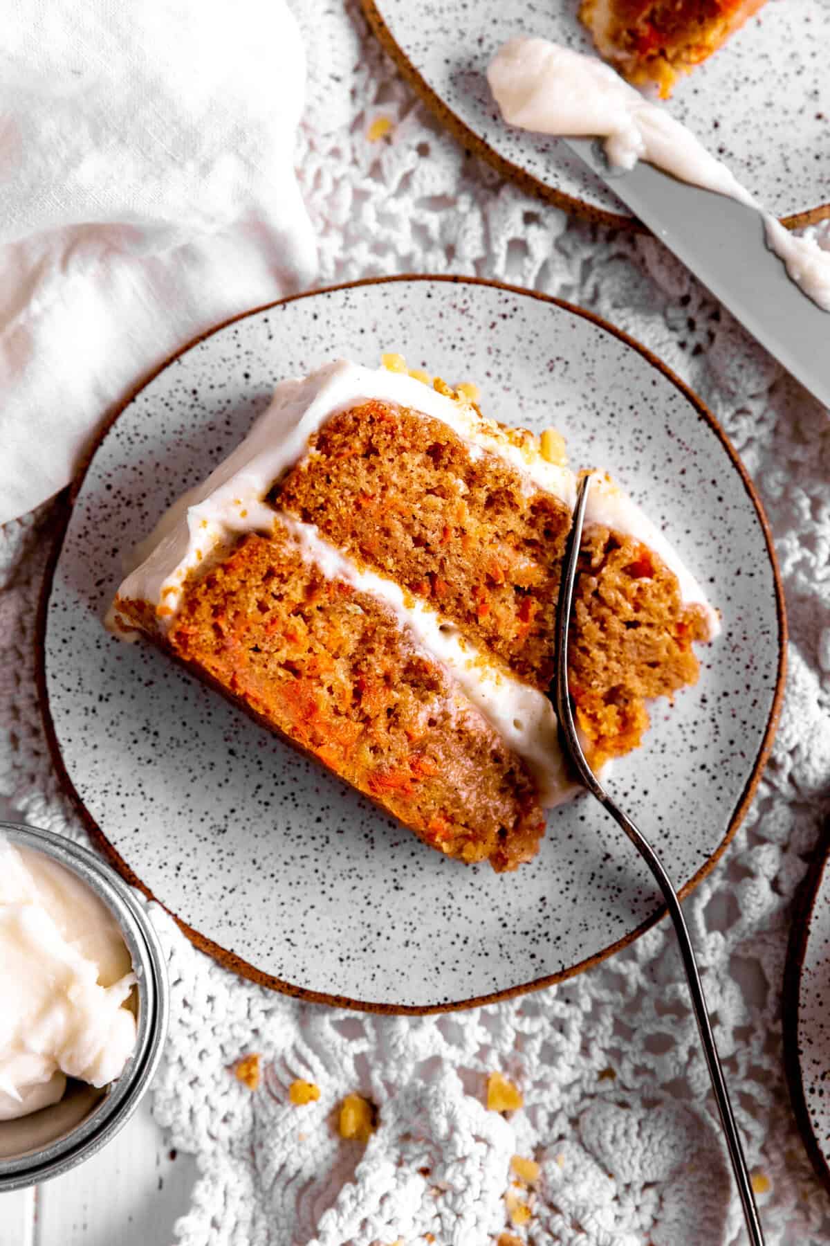 Fork cutting into a slice of carrot cake and a cup of frosting.