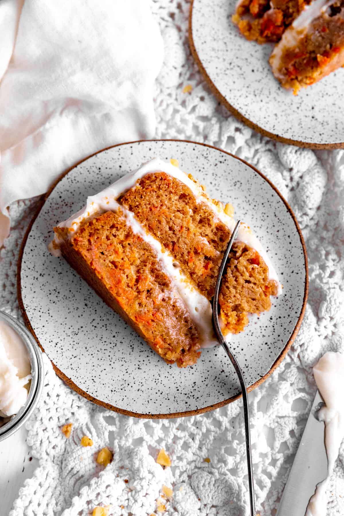 Fork cutting into a slice of carrot cake and a cup of frosting.