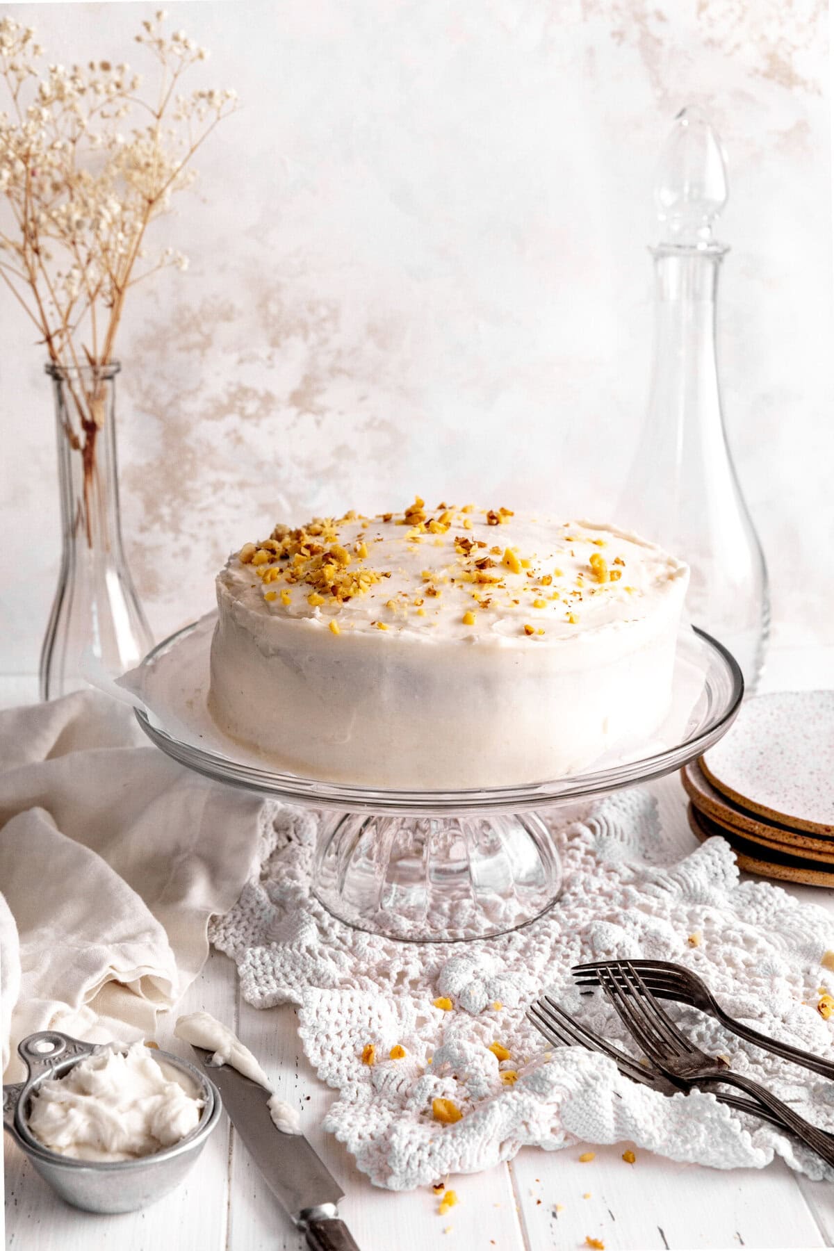 Frosted carrot cake on a glass cake stand, a stack of plates and forks.