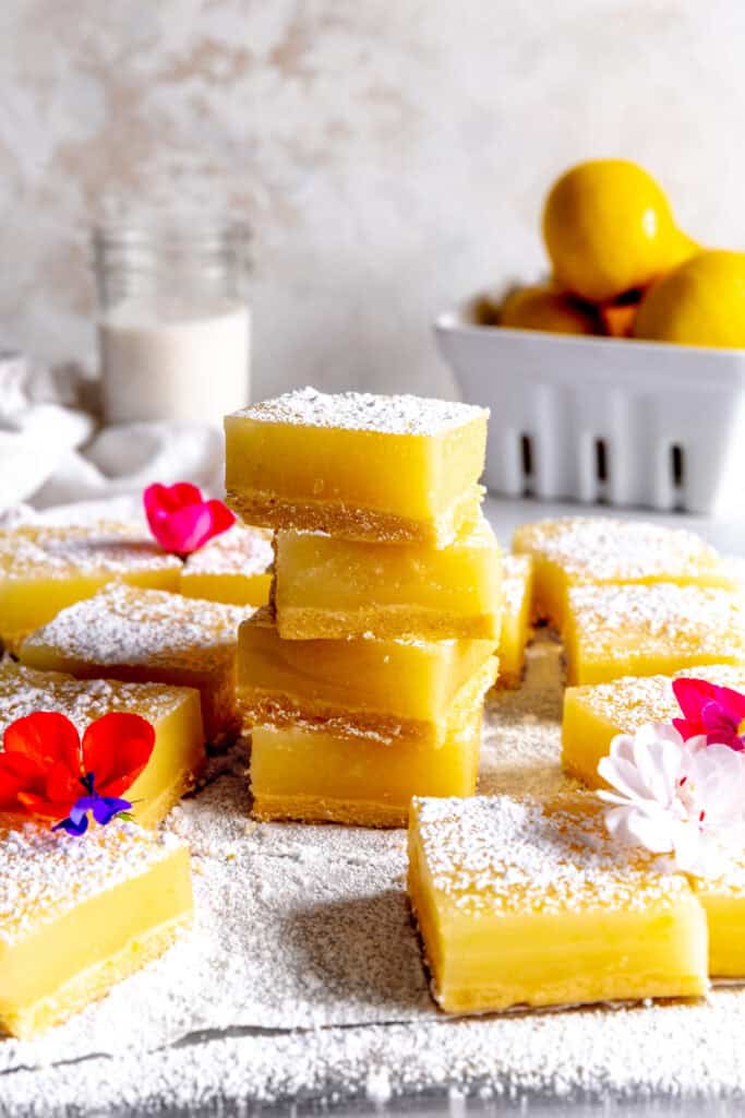 Stack of lemon bars in front of a basket of lemons and a glass of milk.
