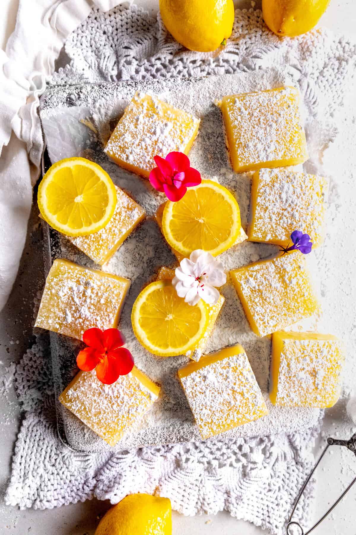 Lemon bars topped with powdered sugar and lemon slices, surrounded by whole lemons.