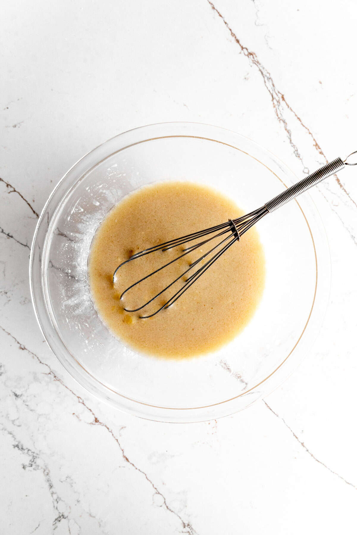 Glass bowl with wet banana cake ingredients and a wire whisk.