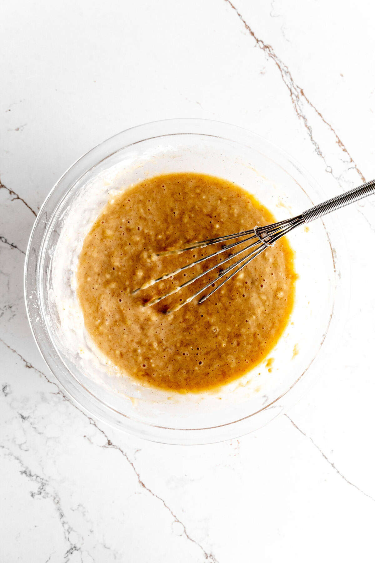 Glass bowl full of banana cake batter and a whisk.