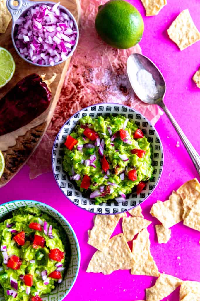 Two bowls of guacamole, a lime, a chili pepper and scattered tortilla chips.