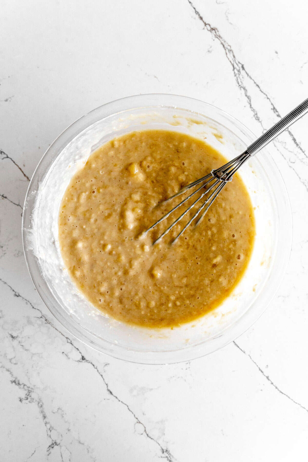 Glass bowl of banana cake batter with a whisk.