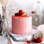 Pink cake with strawberries on top, a jug of milk and a cup of strawberries.