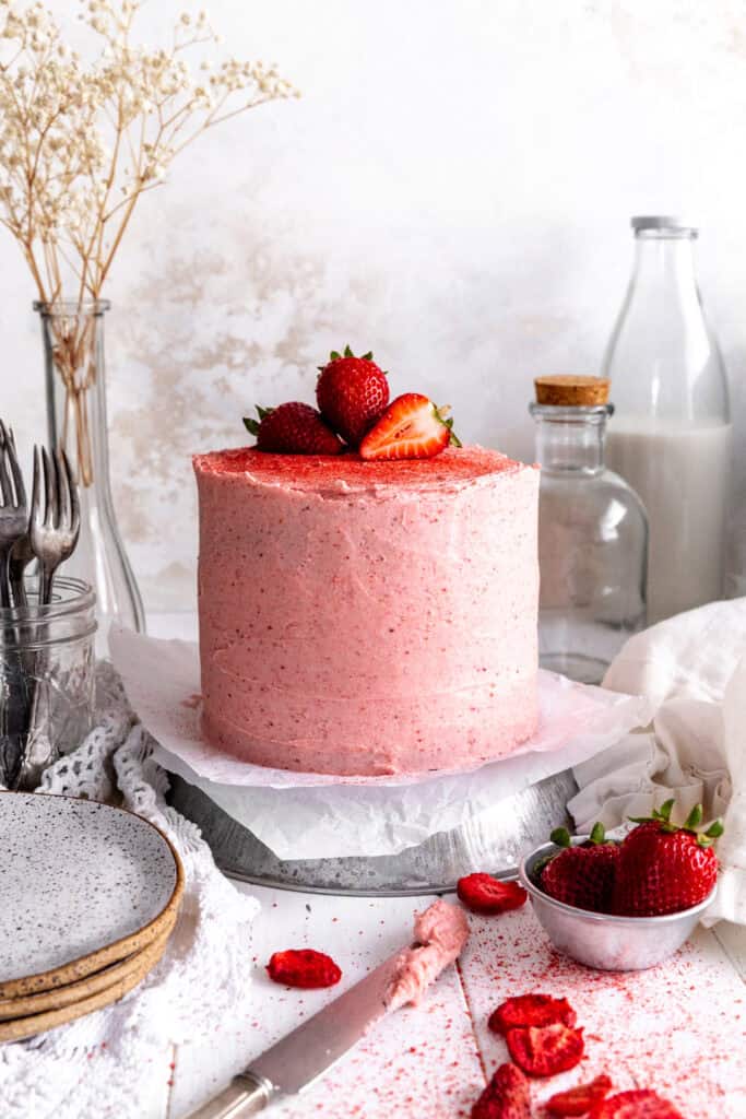 Pink cake with strawberries on top, a jug of milk and a cup of strawberries.